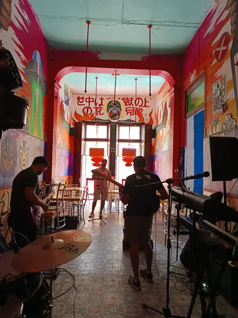Músicos instalando batería y teclado en el salón largo con muros rojos y murales