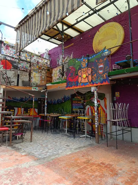 Vista amplia del patio bajo un toldo a rayas, con mesas, bancos de colores y murales