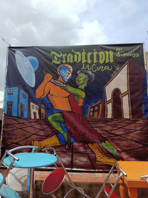 Manta pintada con la frase Tradición del domingo y un luchador bailando, junto a bancos de colores