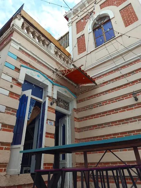 Costado de la casona con puertas azules y una mesa alta turquesa bajo series de focos