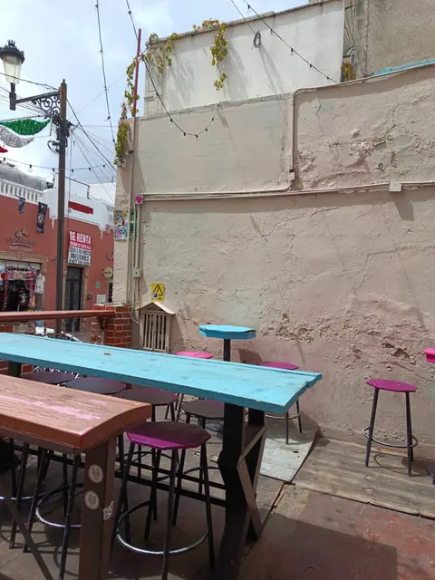 Terraza con mesas de madera turquesa y rosa, bancos altos y un farol de la calle